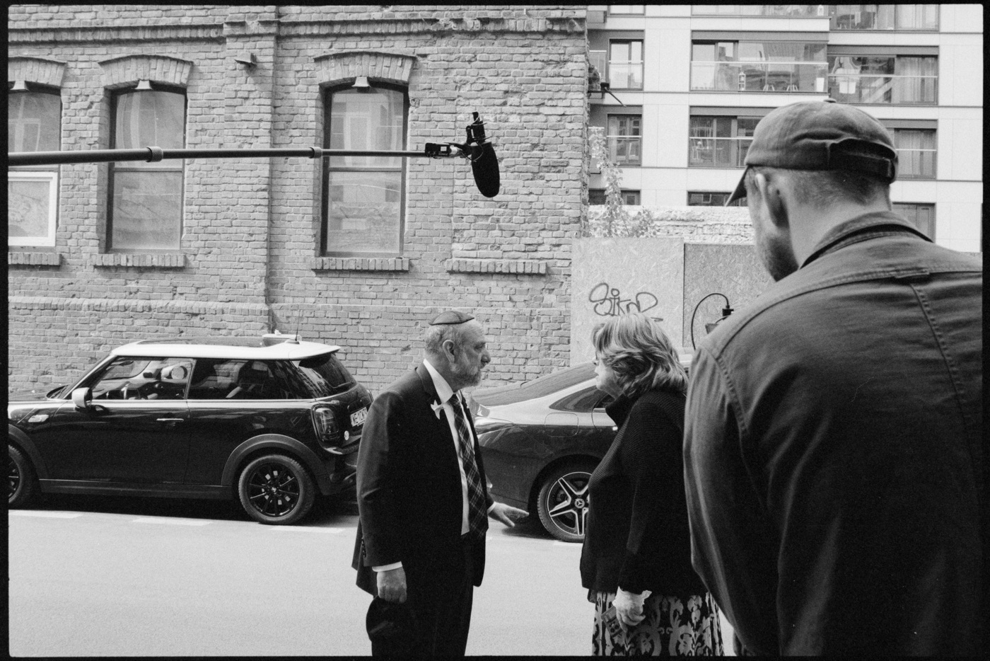 The boom microphone reaches over a street conversation between Ruth and a companion at a brick wall.